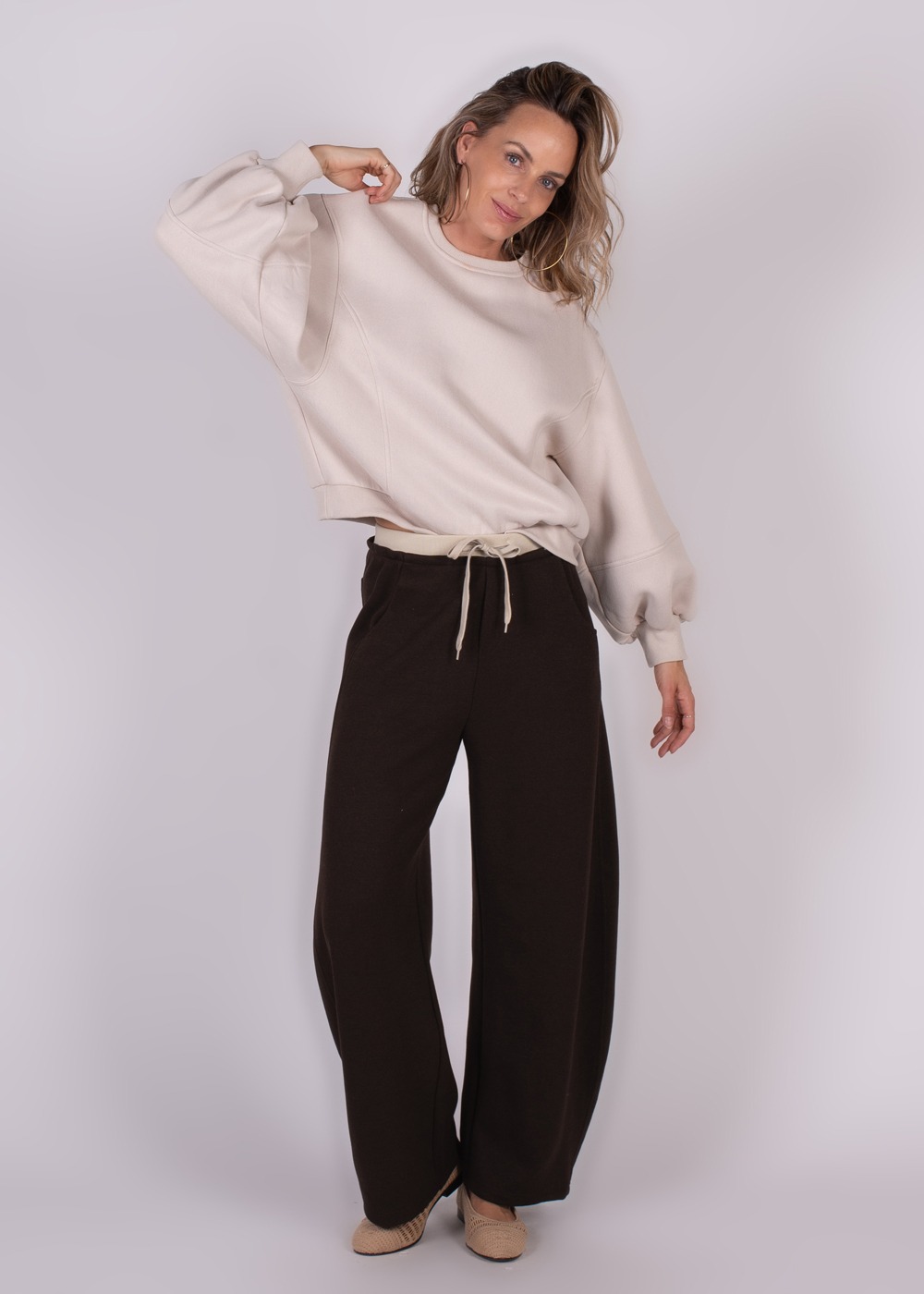 Vrouwelijk model draagt een beige Capri oversized sweater met wijde bruine broek, staand tegen een lichtgrijze achtergrond.