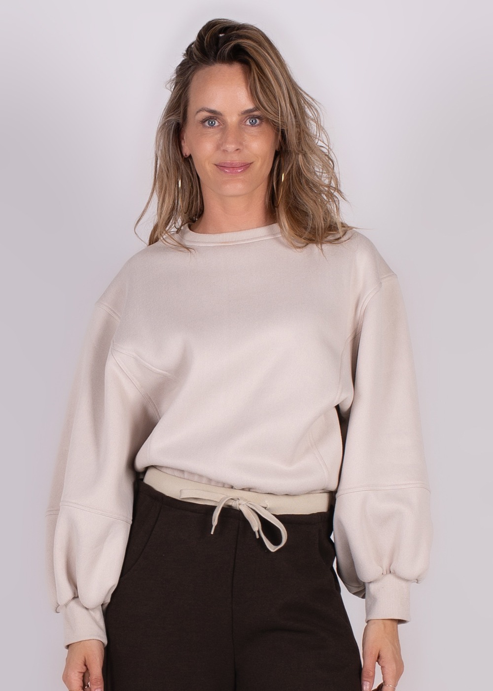 Model draagt een Capri oversized sweater in lichtbeige met lange mouwen en trekt aan koord in donkere broek.