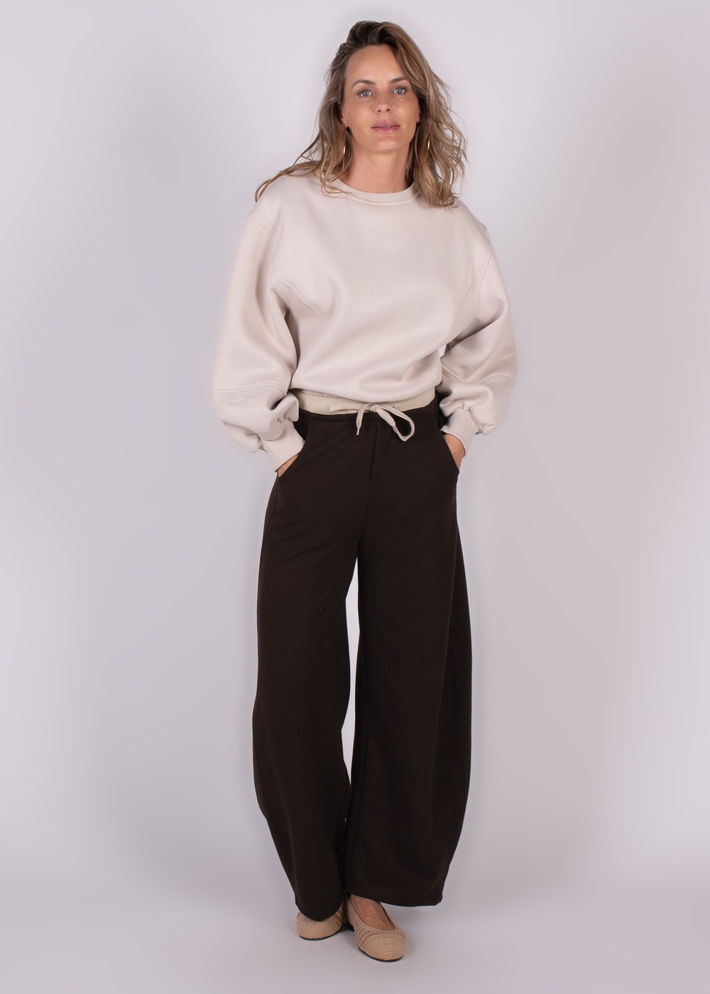 Model draagt een crème oversized sweater genaamd Capri, gecombineerd met losse zwarte broek en lichtbruine schoenen, tegen een neutrale achtergrond.