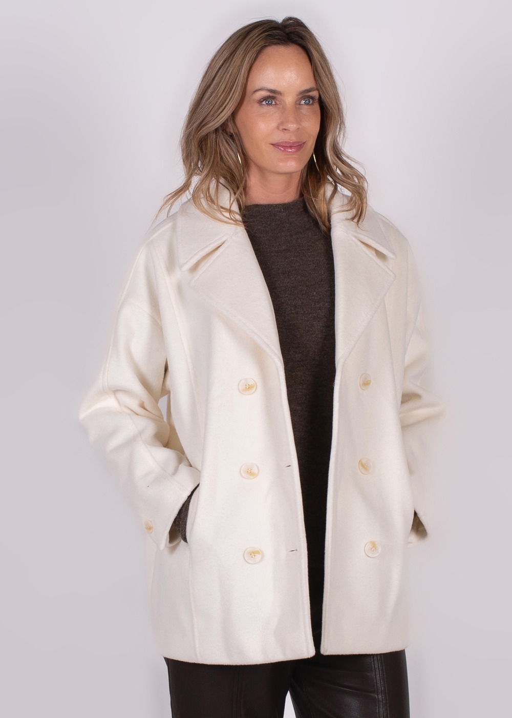 Een vrouw draagt een crèmekleurige, dubbelknoopsluiting mantel genaamd "Depok coat" en poseert met haar handen in de zakken.