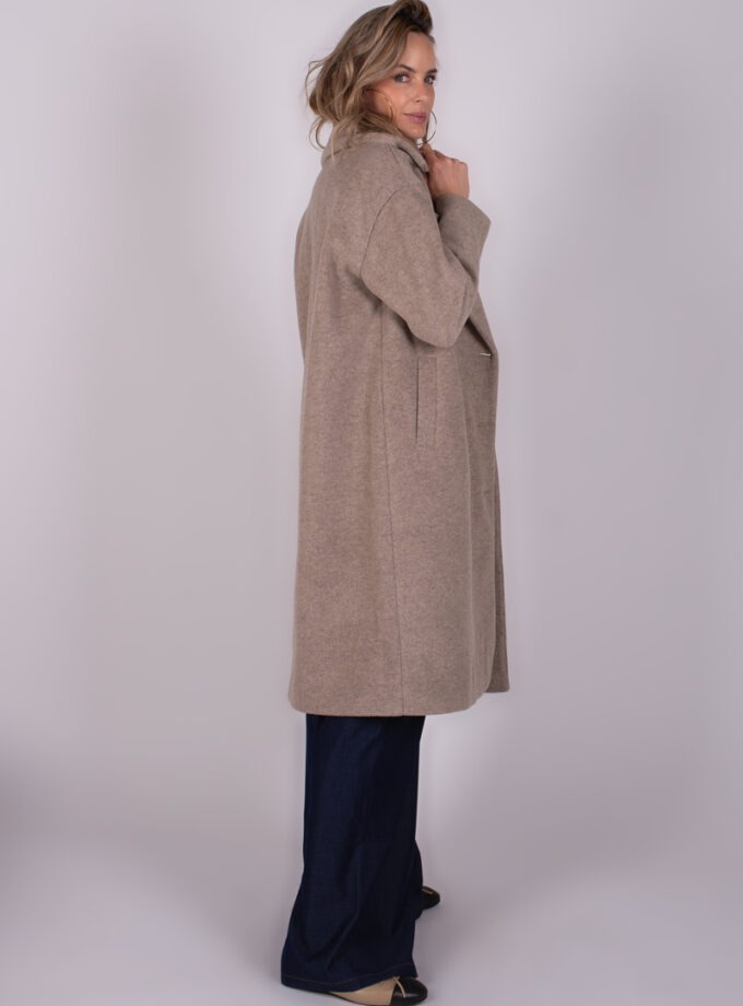 Een vrouw draagt een lange, beige jas genaamd "Depok long coa-227" terwijl ze in profiel richting de camera kijkt.
