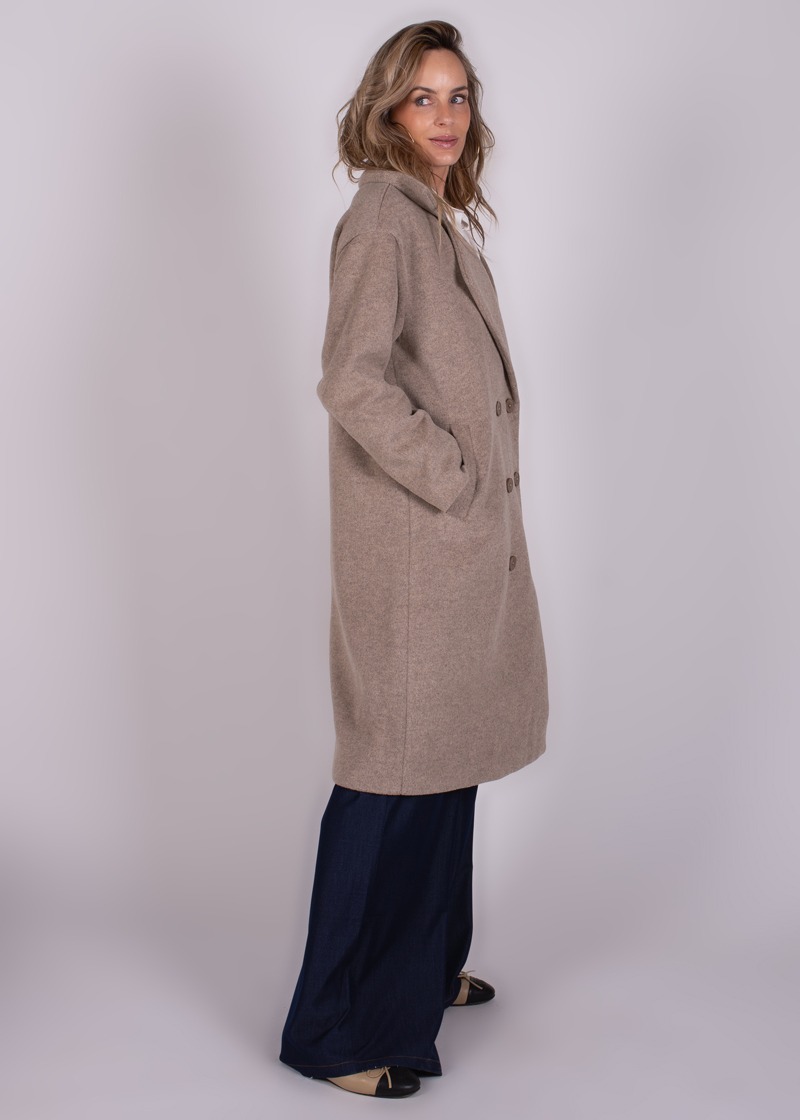 Model draagt een lange, beige jas genaamd "Depok long coa-229" met de handen in de zakken, gekleed in donkere broek en lichte schoenen, gefotografeerd van de zijkant.