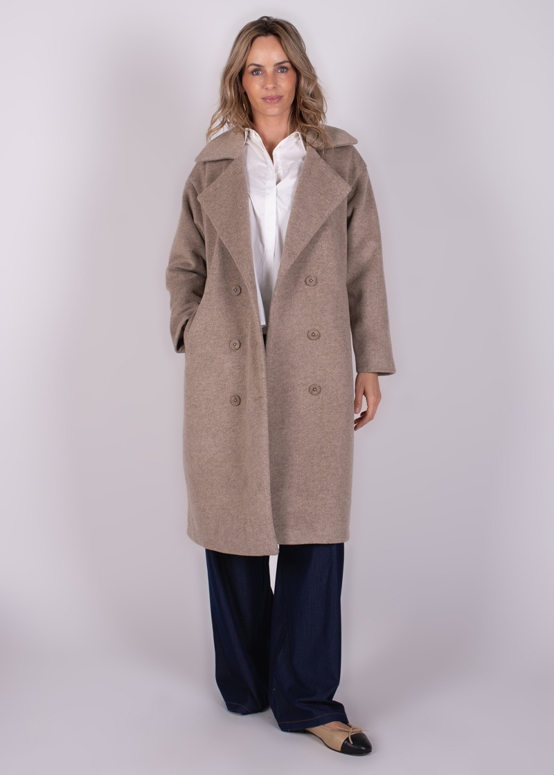 Een vrouw poseert in een camel-kleurige lange jas genaamd "Depok" over een witte blouse en donkerblauwe broek, op een neutrale achtergrond.