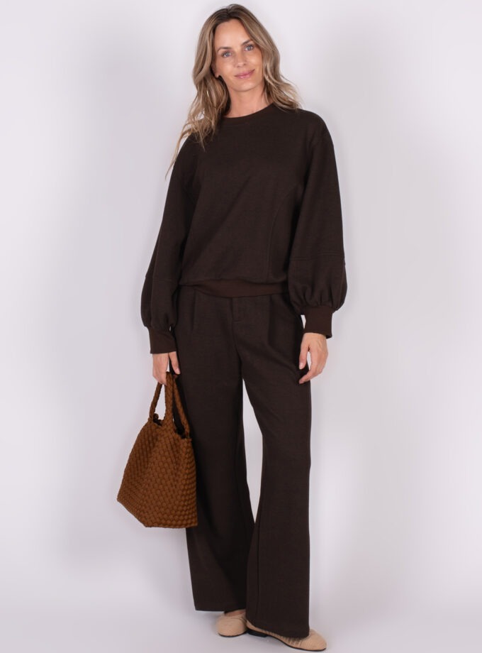 Een vrouw poseert met een bruine oversize sweater en bijpassende broek, terwijl ze een geweven bruine tas vasthoudt.