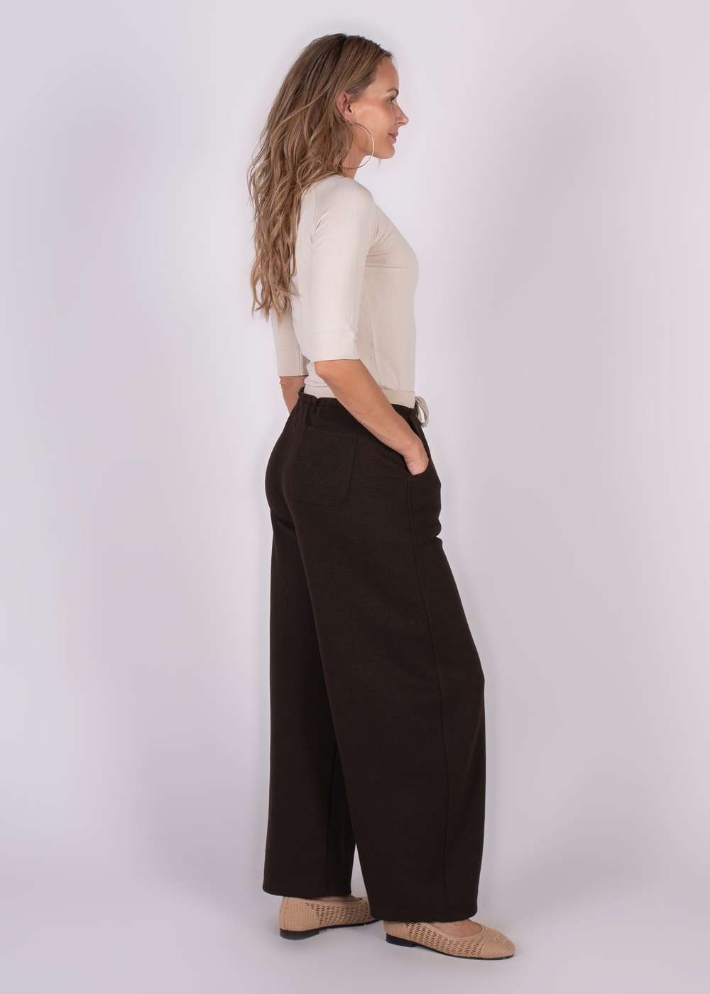 Een vrouw poseert in een zijaanzicht met een crème top en een bruine Helsinki pantalon.