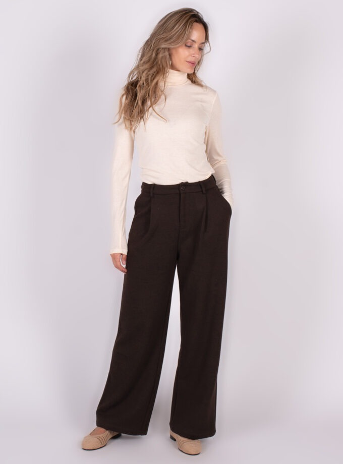 Model draagt wijde donkere broek met hoge taille en lichte coltrui, poserend tegen een neutrale achtergrond.