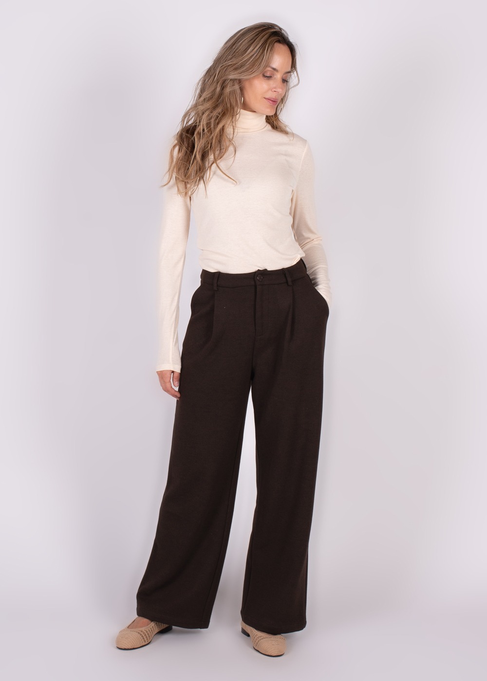 Model draagt wijde donkere broek met hoge taille en lichte coltrui, poserend tegen een neutrale achtergrond.