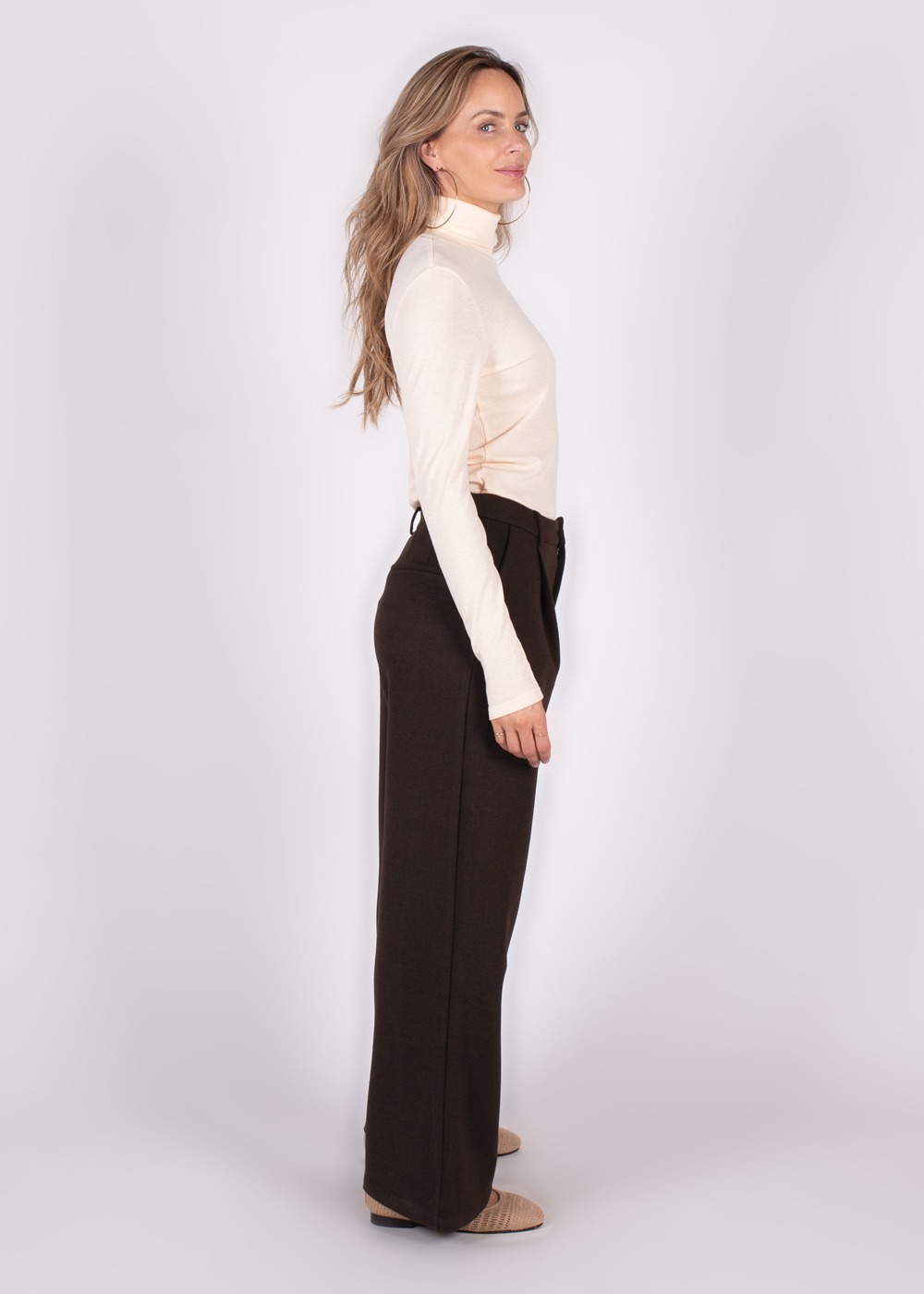 Een vrouw met lang haar poseert in een crème coltrui en bruine "Helsinki piran pants", gefotografeerd van de zijkant tegen een witte achtergrond.