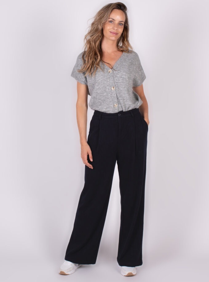 Model draagt een grijze top met knopen en zwarte wijde broek, gefotografeerd tegen een lichte achtergrond.