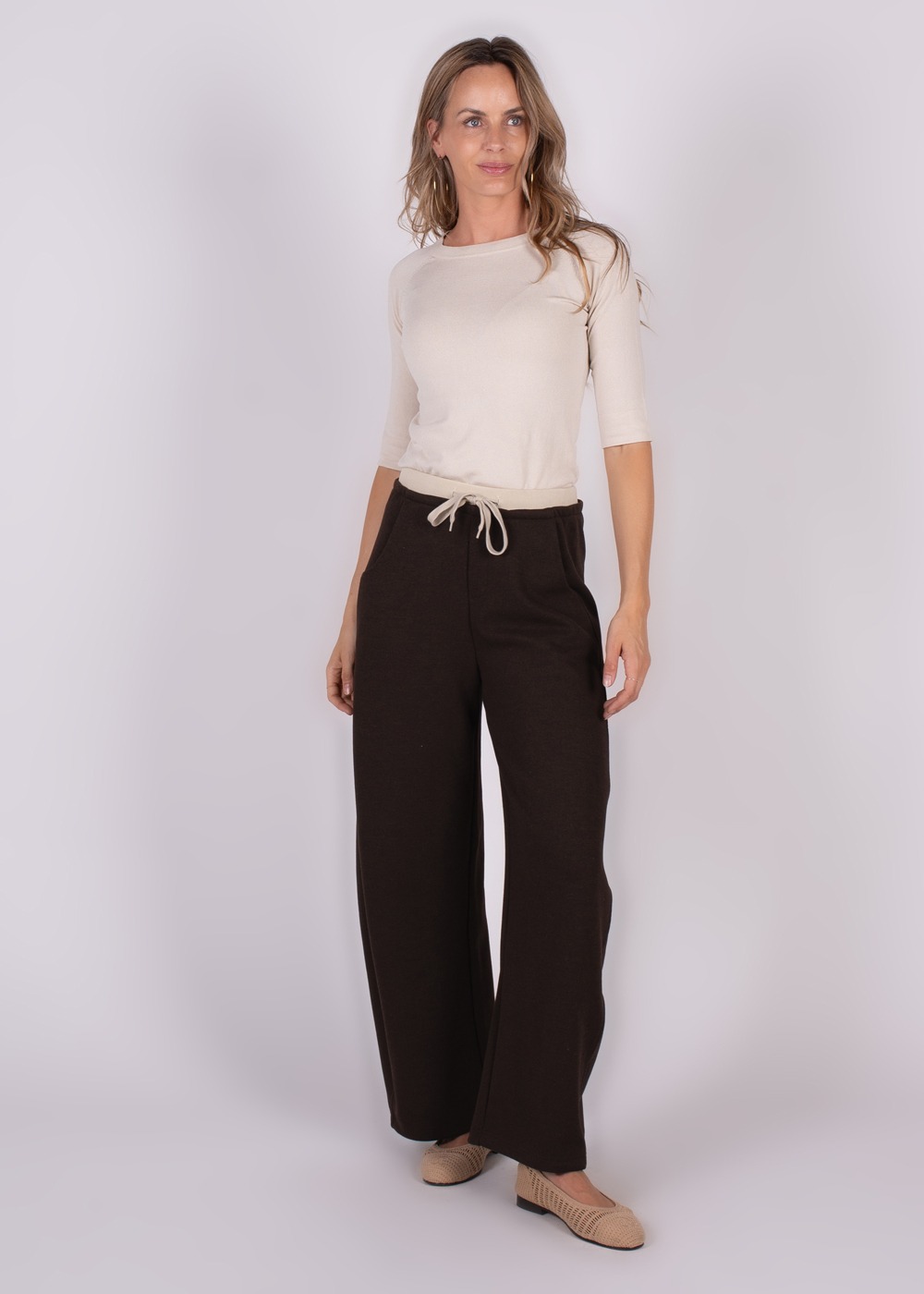 Vrouwelijk model draagt een crèmekleurige top en een donkere Helskini pantalon met wijde pijpen, afgebeeld op een eenvoudige achtergrond.