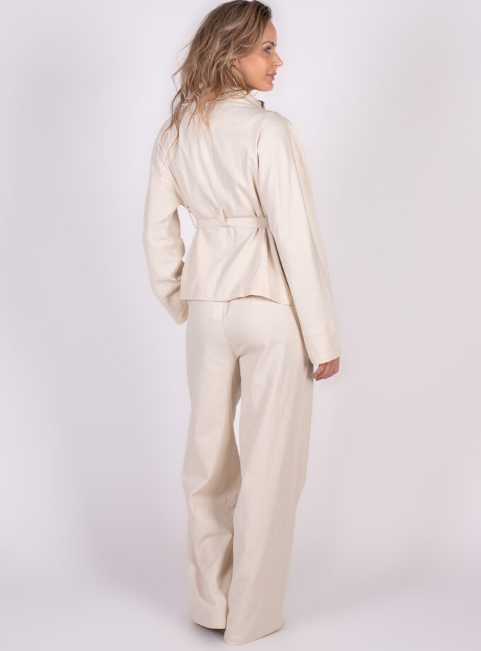 Vrouw in een lichte, stijlvolle broek en bijpassende blazer, gefotografeerd van opzij tegen een neutrale achtergrond.