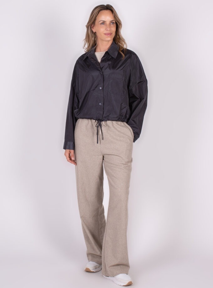 Een vrouw draagt een zwarte Lubia blouse en lichte wijde broek, met witte sneakers, gefotografeerd vanuit een frontaal perspectief tegen een witte achtergrond.