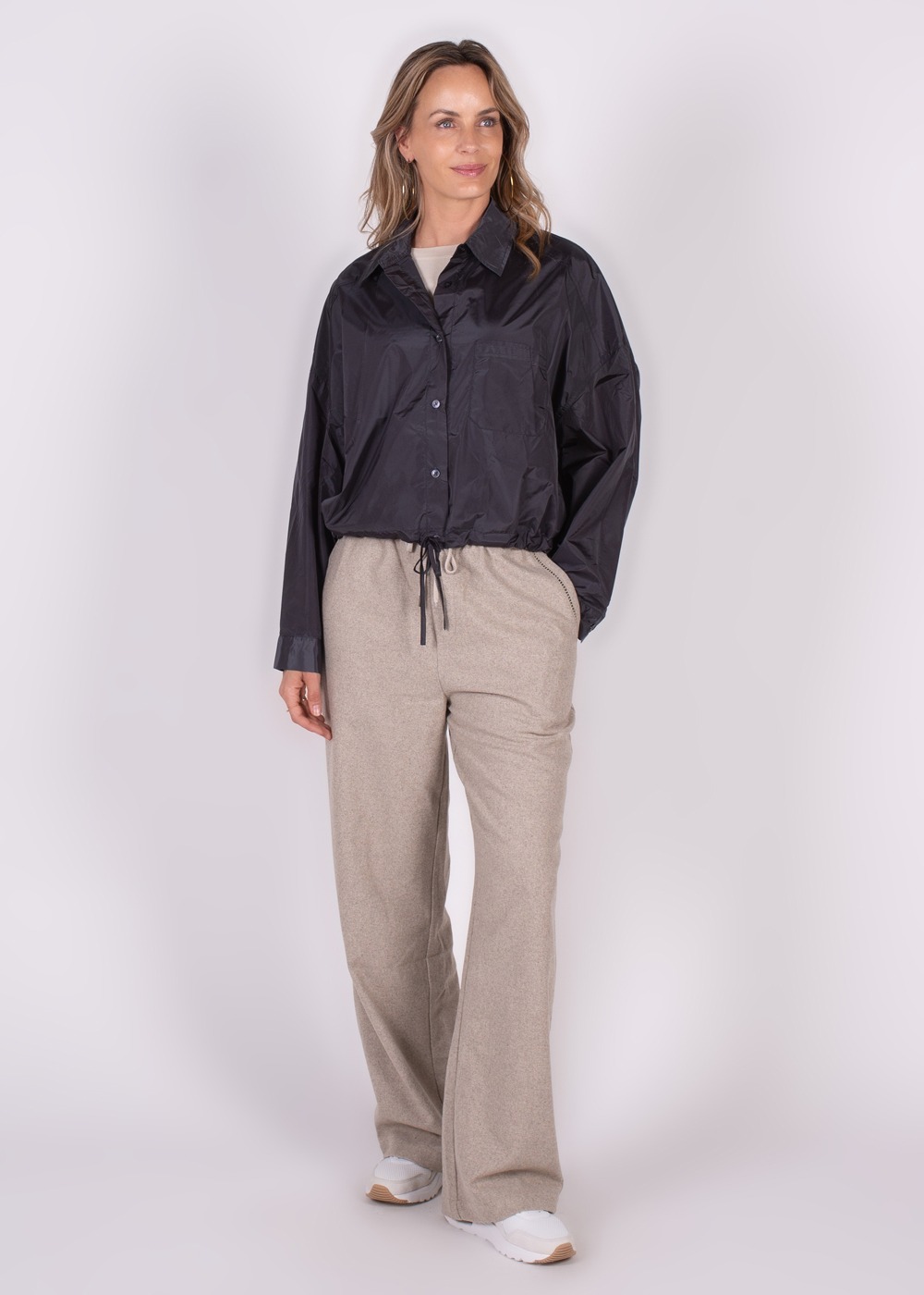 Een vrouw draagt een zwarte Lubia blouse en lichte wijde broek, met witte sneakers, gefotografeerd vanuit een frontaal perspectief tegen een witte achtergrond.