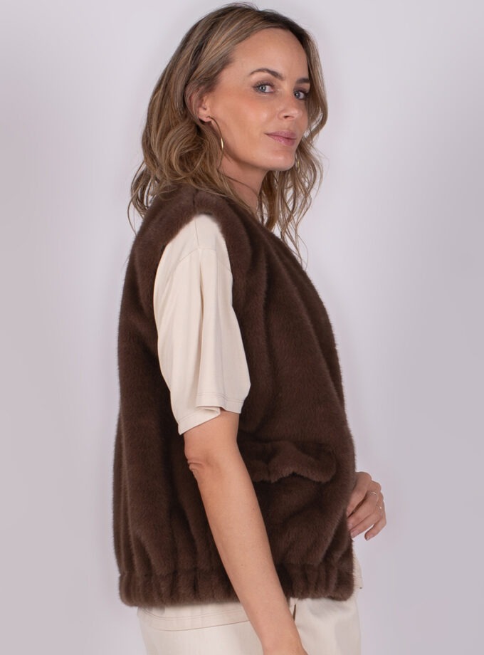 Model draagt een bruin bontvest over een korte mouwen T-shirt, zijwaarts gefotografeerd tegen een neutrale achtergrond.