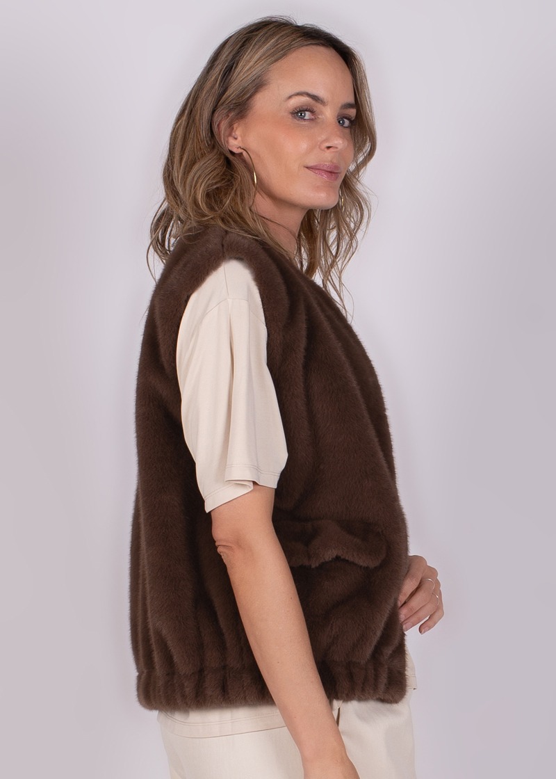 Model draagt een bruin bontvest over een korte mouwen T-shirt, zijwaarts gefotografeerd tegen een neutrale achtergrond.