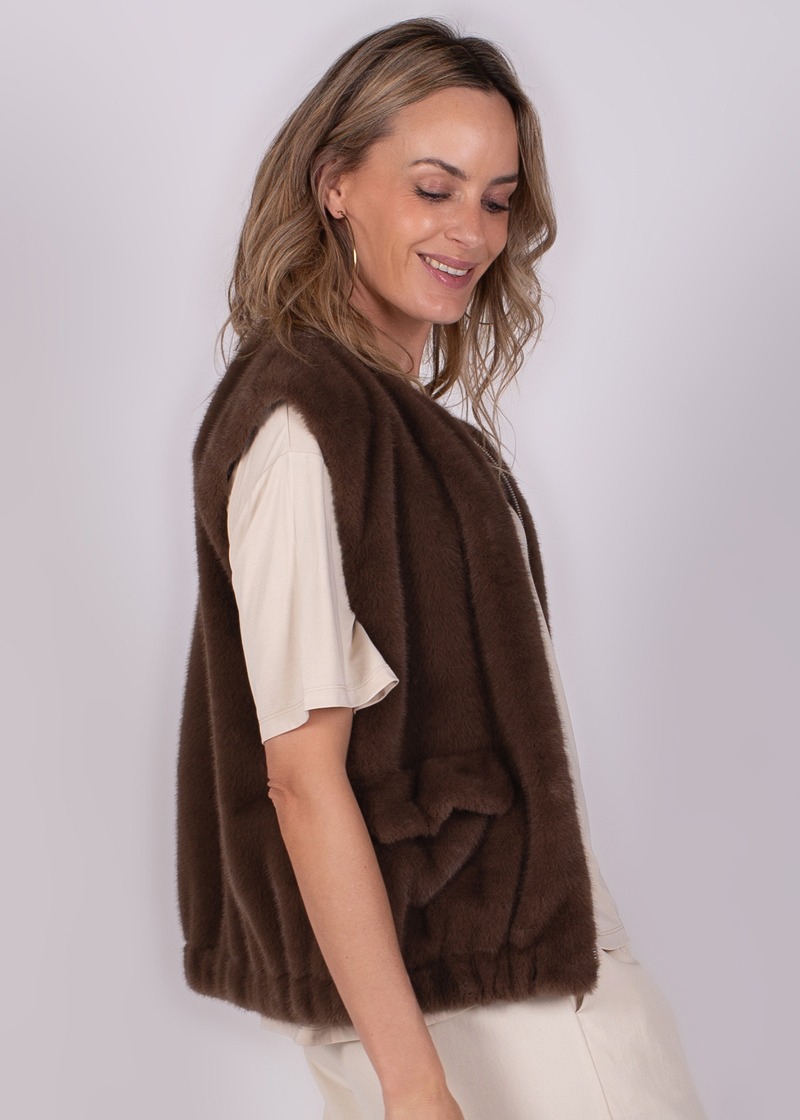 Model draagt een bruin mouwloos vest over een beige T-shirt, kijkend naar beneden met een glimlach in zijaanzicht.