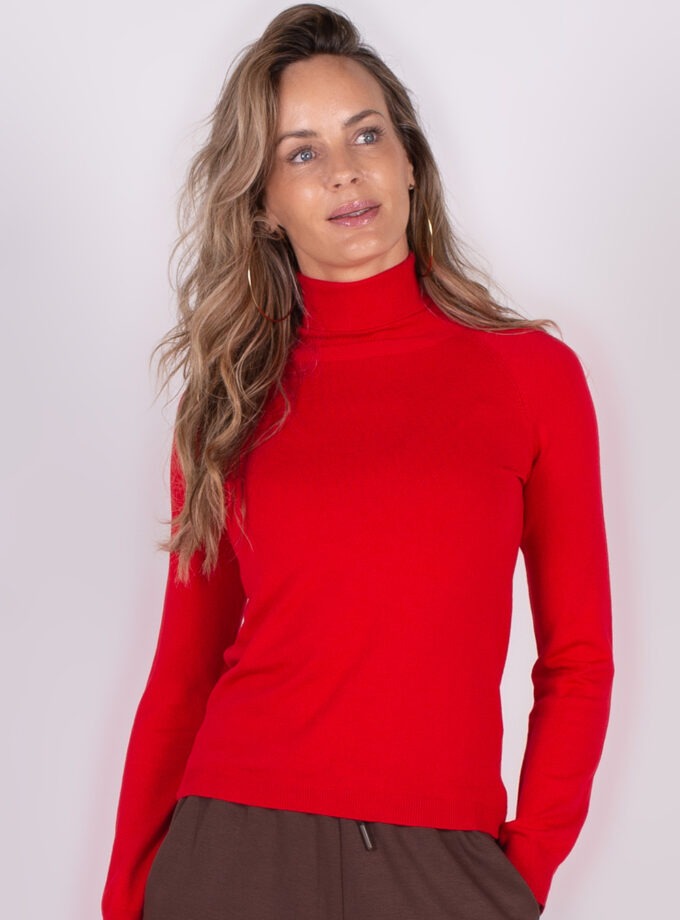Een persoon draagt een felrode coltrui gecombineerd met bruine broek, tegen een neutrale achtergrond.