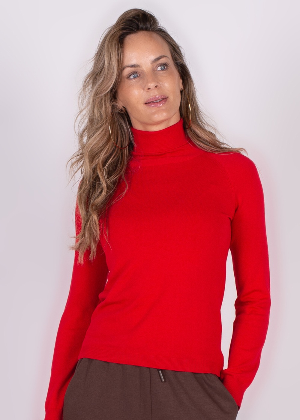 Een persoon draagt een felrode coltrui gecombineerd met bruine broek, tegen een neutrale achtergrond.