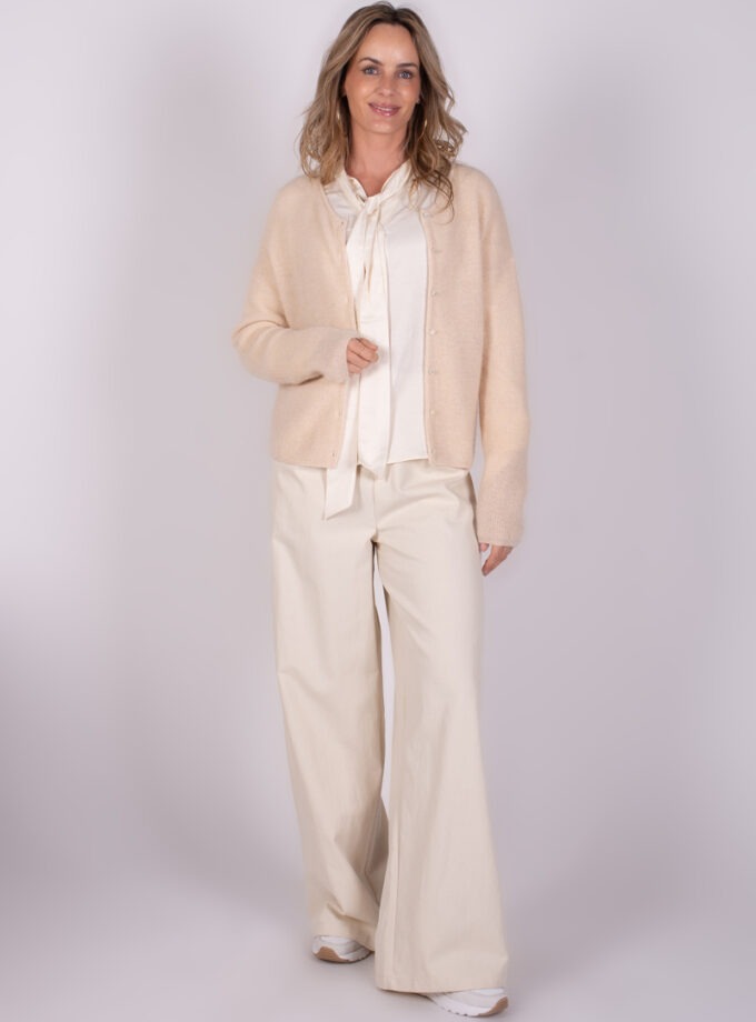 Een vrouw draagt een Narvik cardigan in een lichte kleur, gecombineerd met een witte blouse en losse, lichtgekleurde broek, staand tegen een effen achtergrond.