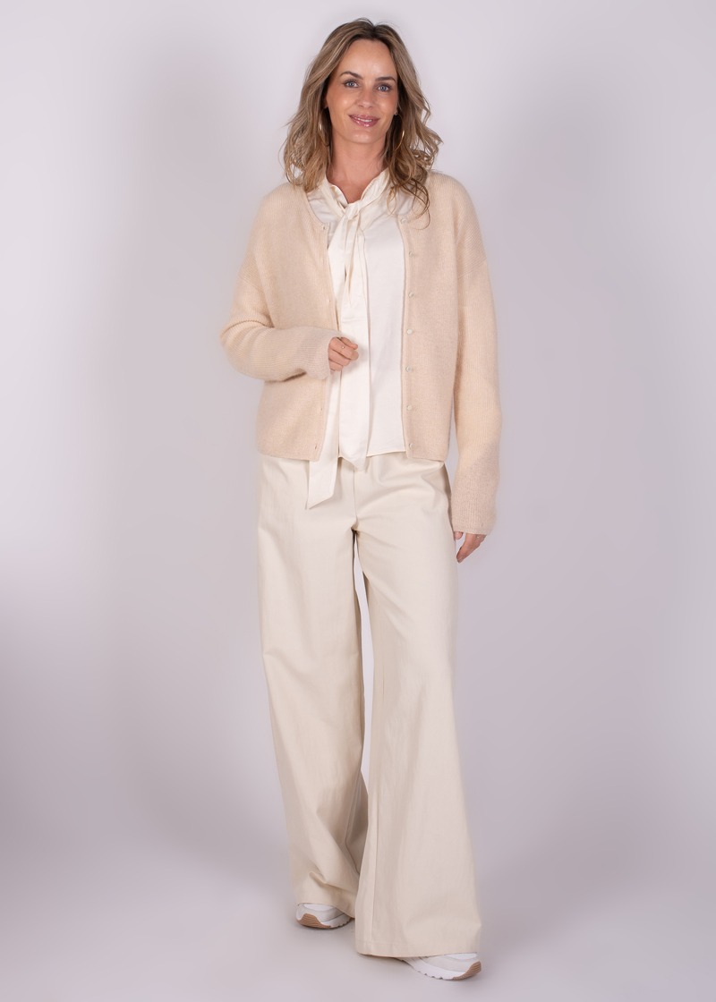 Een vrouw draagt een Narvik cardigan in een lichte kleur, gecombineerd met een witte blouse en losse, lichtgekleurde broek, staand tegen een effen achtergrond.