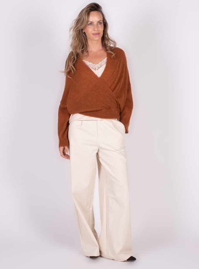 Een vrouw poseert in een warme bruine overslag cardigan en wijde crèmekleurige broek voor een neutrale achtergrond.