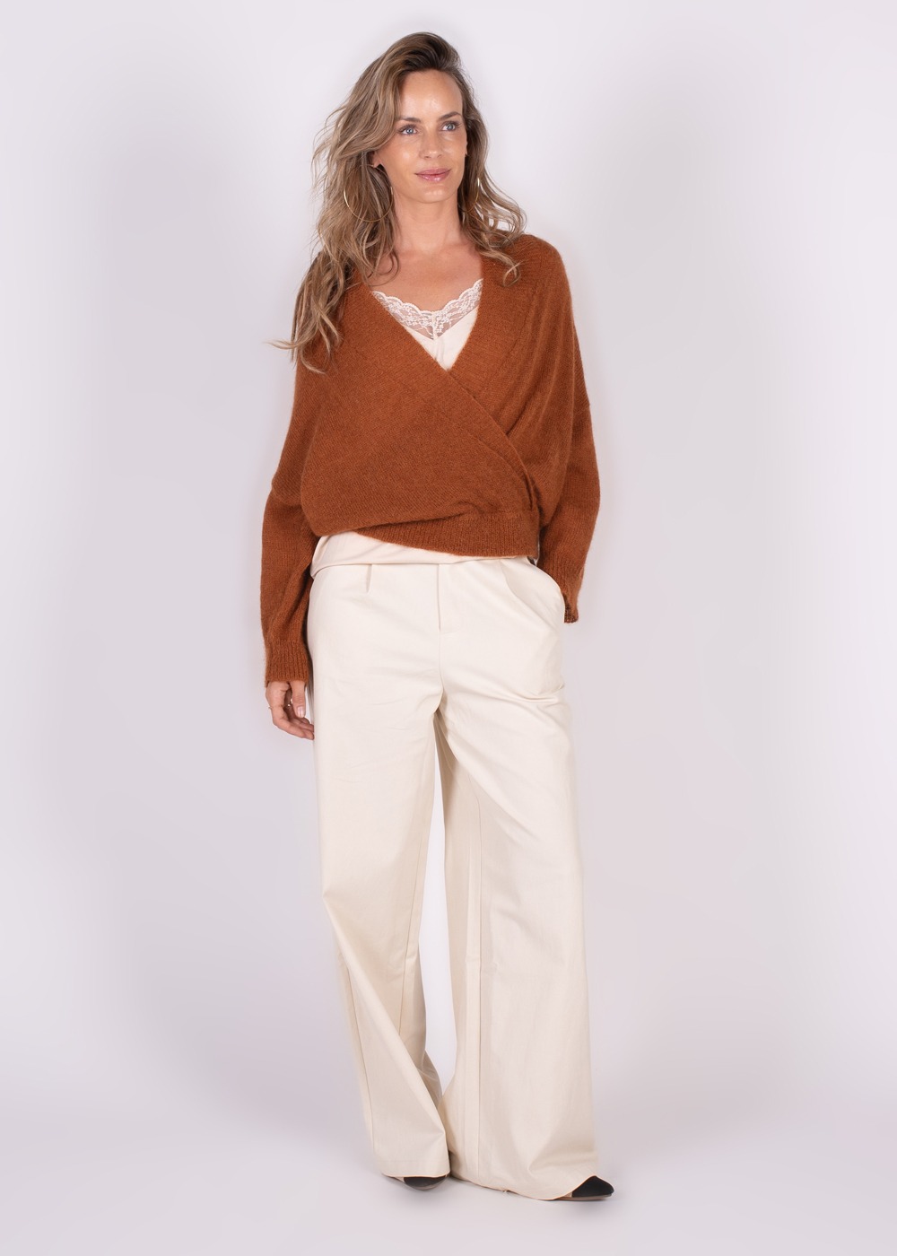 Een vrouw poseert in een warme bruine overslag cardigan en wijde crèmekleurige broek voor een neutrale achtergrond.