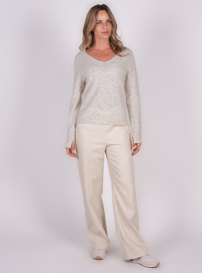 Vrouw in een lichte V-hals trui en wijde broek, staand op een witte achtergrond.