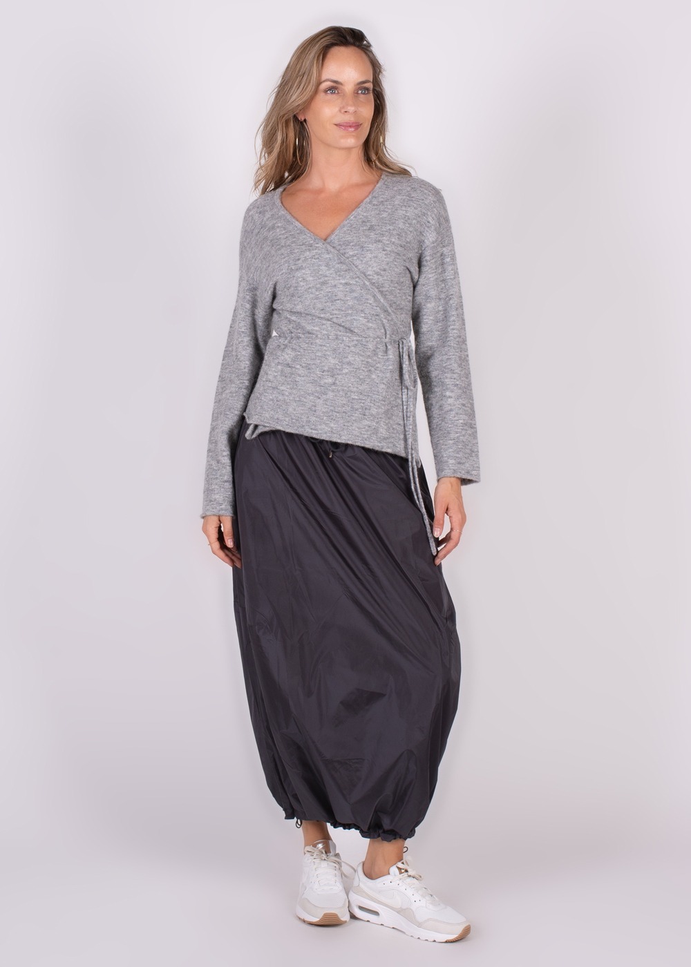 Een vrouw draagt een grijze Odense cardigan met een zwarte rok en witte sneakers, weergegeven in een volledige lichaamshoek.