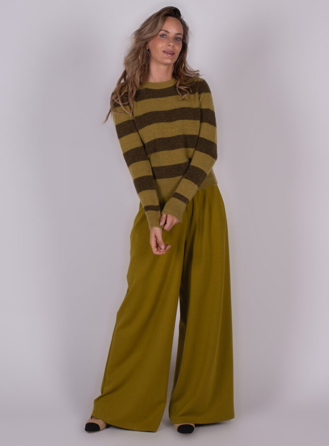 Model draagt een olijfgroen met bruine gestreepte trui ("Odense stripe pullover") en losse groene broek, tegen een neutrale achtergrond.