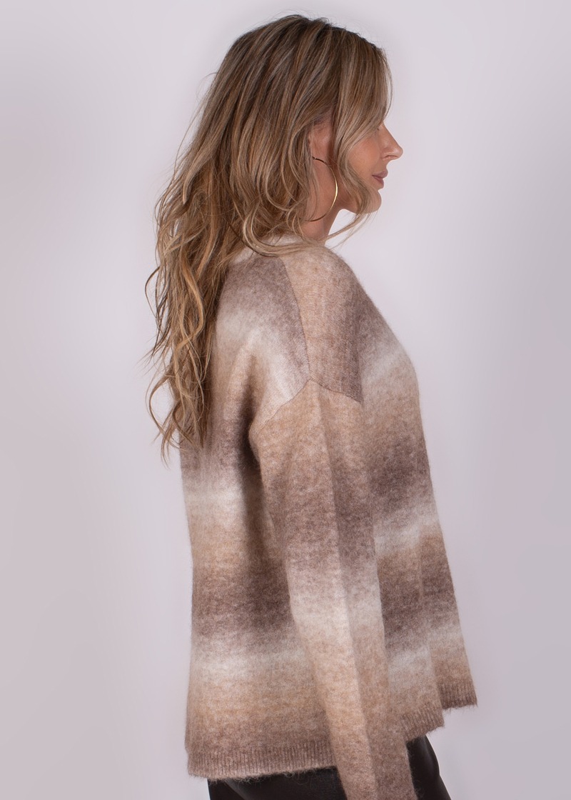 Zijaanzicht van een persoon met lang blond haar die een beige en bruine gebreide trui draagt tegen een grijze achtergrond.