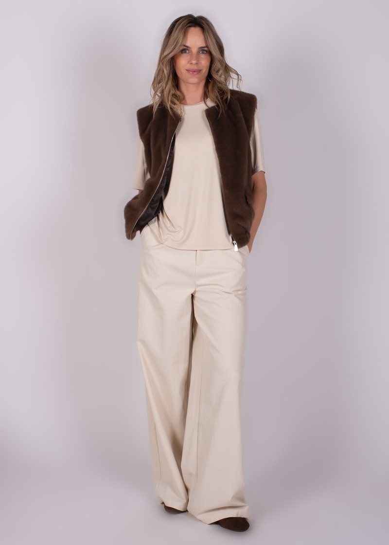 Vrouw met lang blond haar draagt een bruine vest en beige outfit, staand tegen een grijze achtergrond.