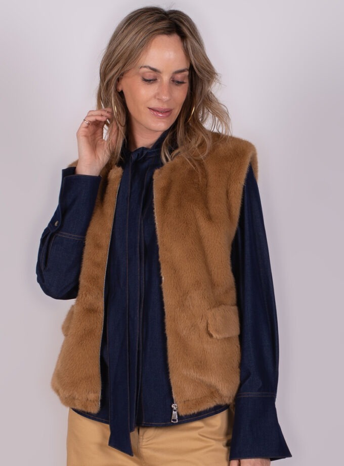 Een vrouw poseert in een stijlvolle bruine bontgilet en een donkerblauwe blouse met wijde mouwen, tegen een neutrale achtergrond.