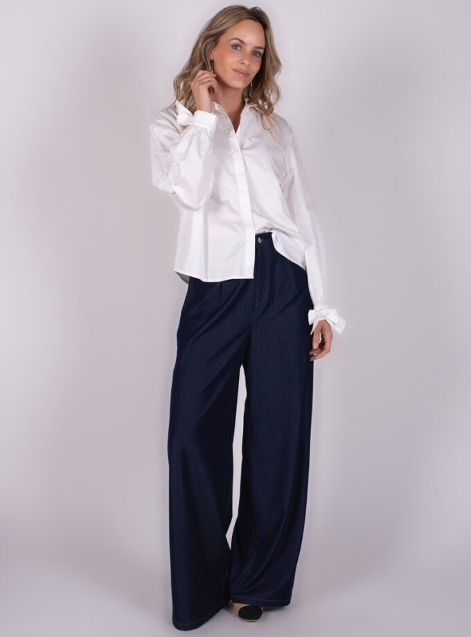 Een persoon die een witte Perth-blouse en donkerblauwe wijde broek draagt, poseert tegen een neutrale achtergrond.