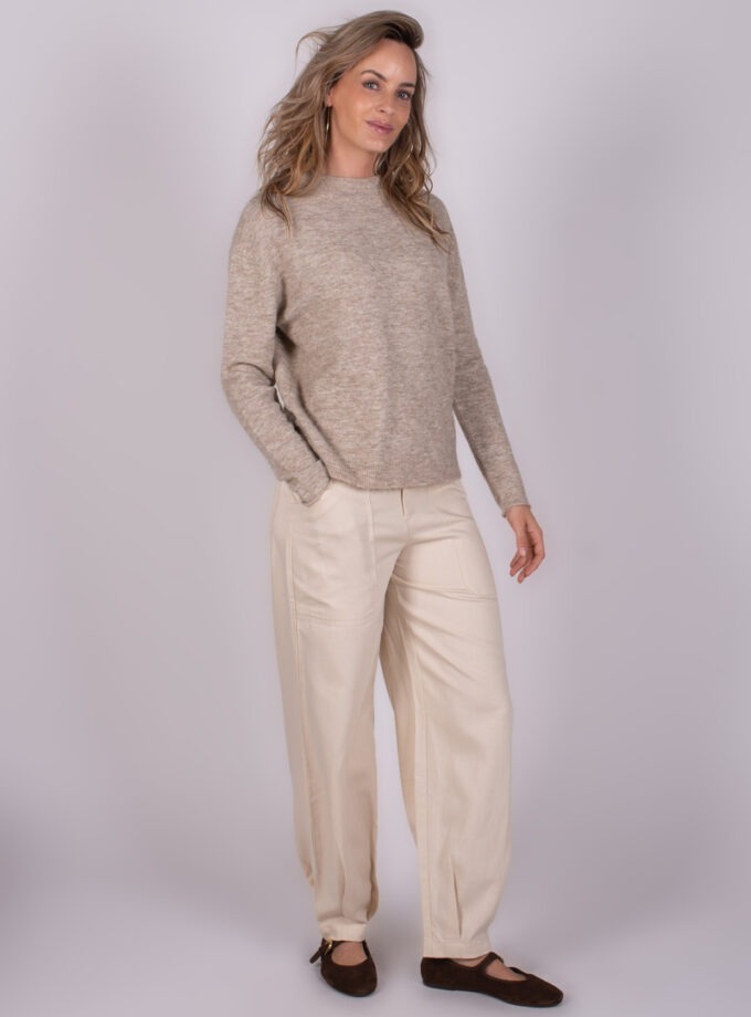 Vrouw in een beige trui en lichte broek, poserend tegen een neutrale achtergrond.