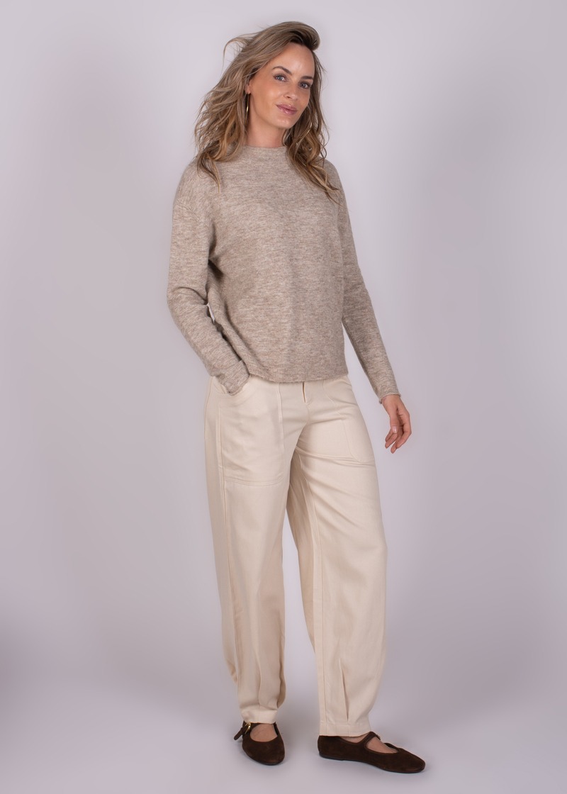 Vrouw in een beige trui en lichte broek, poserend tegen een neutrale achtergrond.