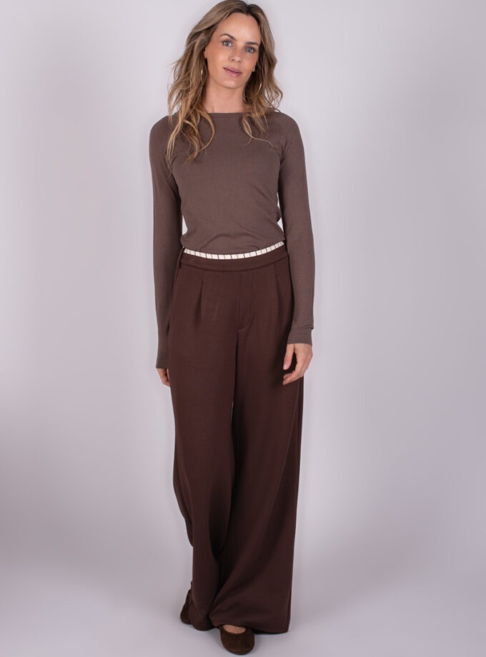 Model in bruine trui en wijde bruine broek, gefotografeerd tegen een grijze achtergrond.