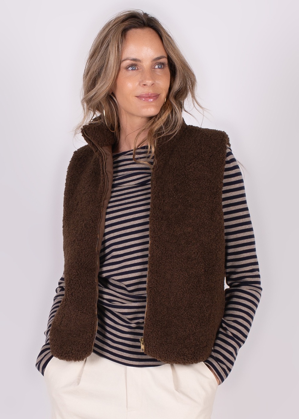 Een vrouw poseert in een bruine Regina Gilet en een gestreepte trui, tegen een lichte achtergrond.