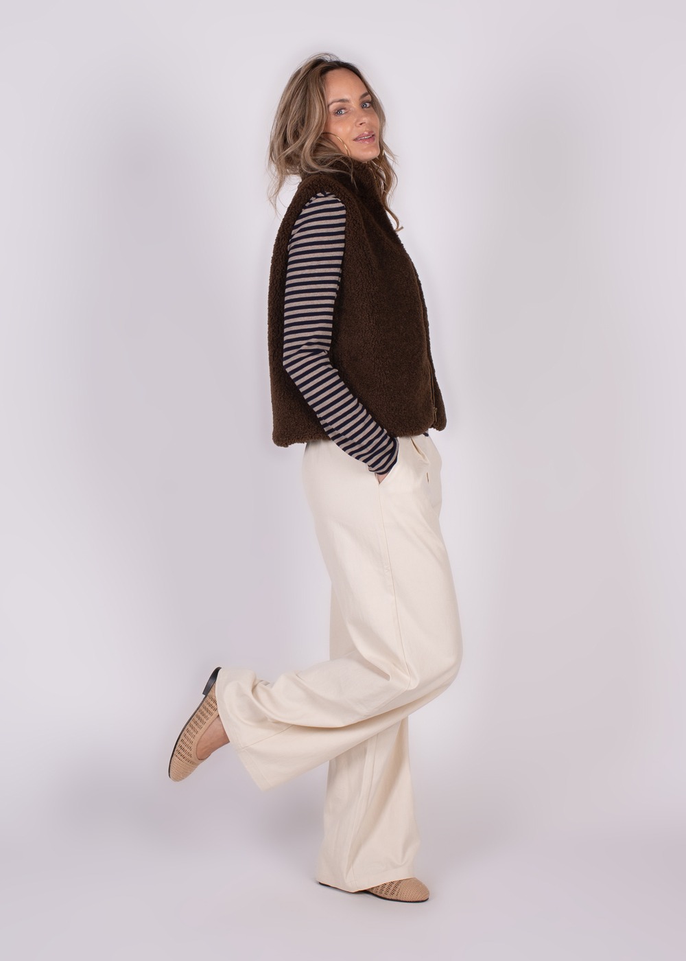 Een vrouw draagt een bruine gilet over een gestreepte trui met witte broek en beige schoenen, kijkend over haar schouder.