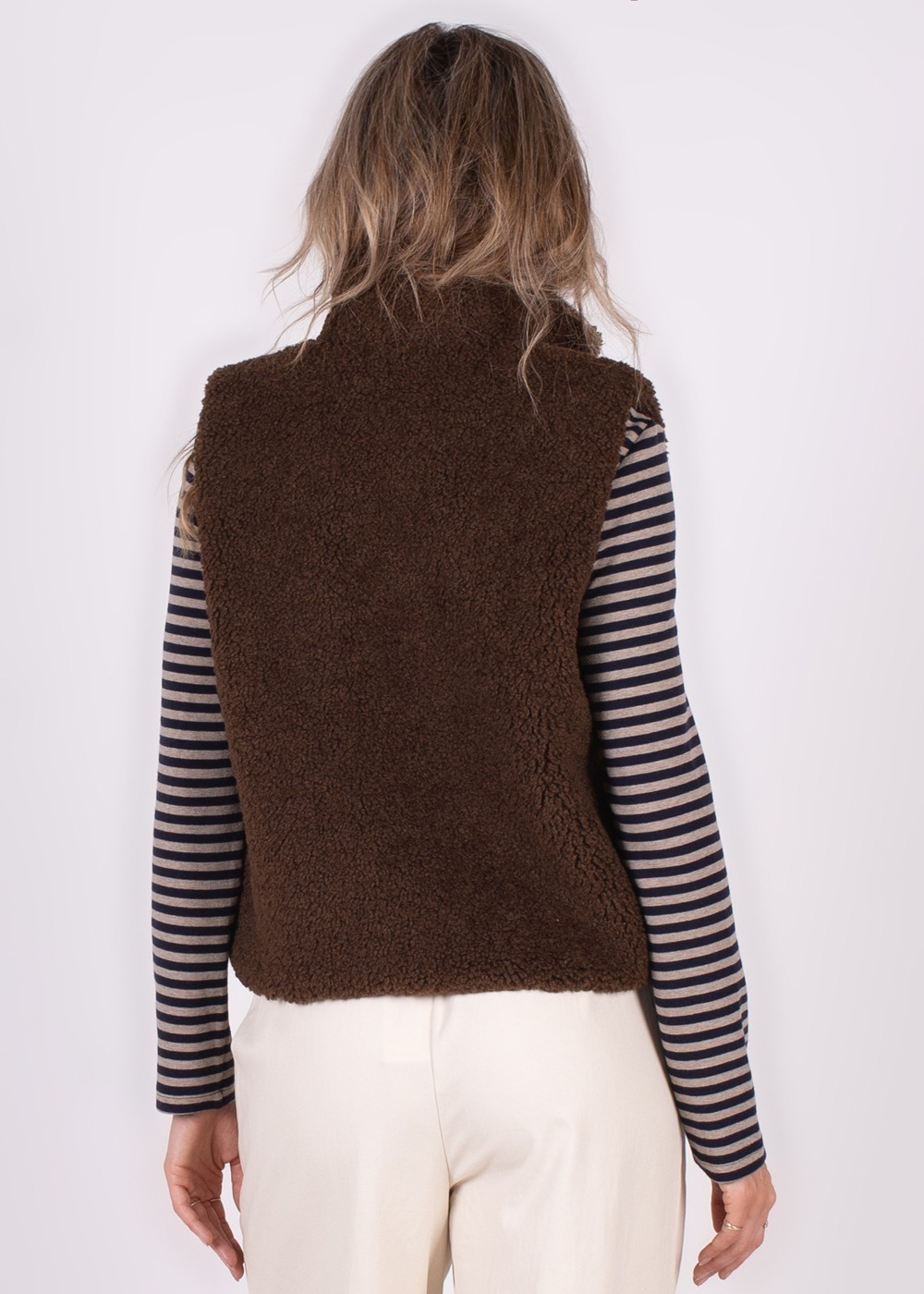 Achteraanzicht van een persoon met lang haar die een bruin Regina Gilet draagt over een gestreepte trui en een witte broek.
