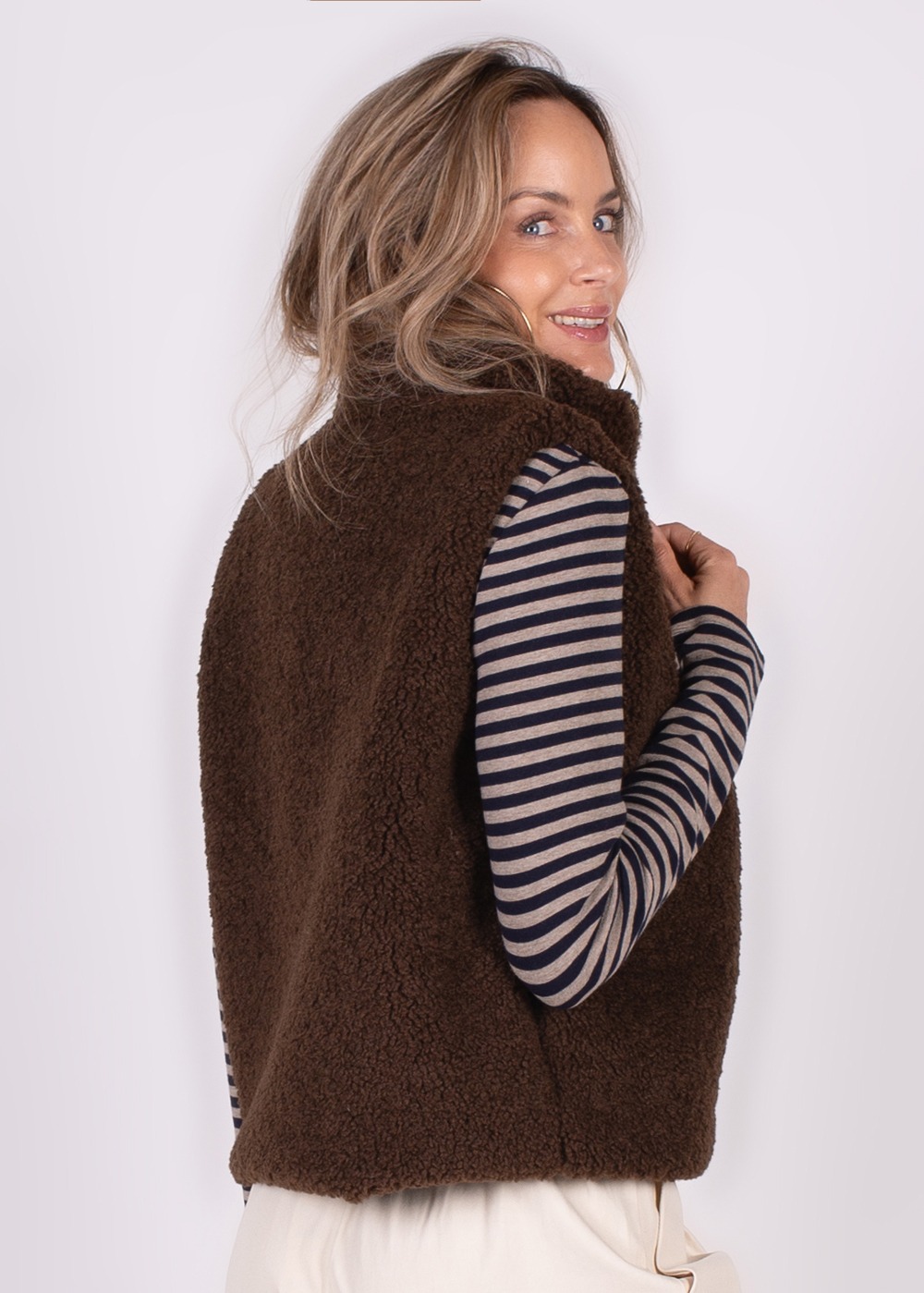 Een vrouw met lang haar draagt een bruin, donzig gilet over een gestreept shirt, kijkend over haar schouder.