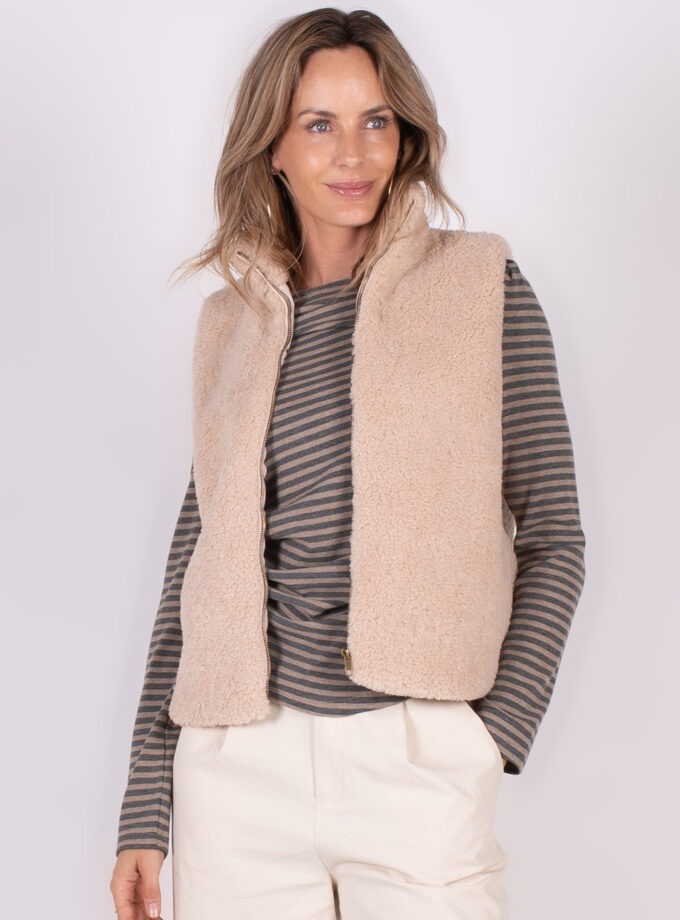Model draagt een lichtroze, wollen gilet over een gestreepte top en lichte broek, staand tegen een neutrale achtergrond.