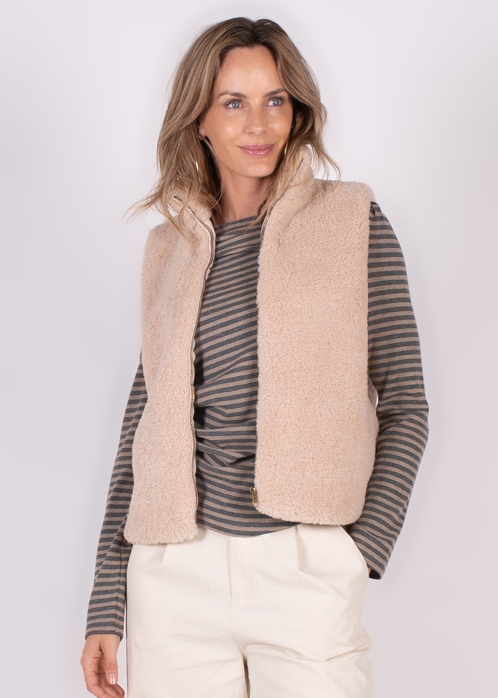 Model draagt een lichtroze, wollen gilet over een gestreepte top en lichte broek, staand tegen een neutrale achtergrond.