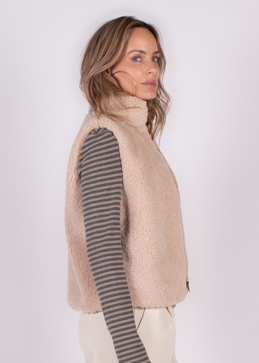 Een vrouw draagt een beige fleecegilet genaamd "Regina" en kijkt zijwaarts, met een gestreepte trui eronder.