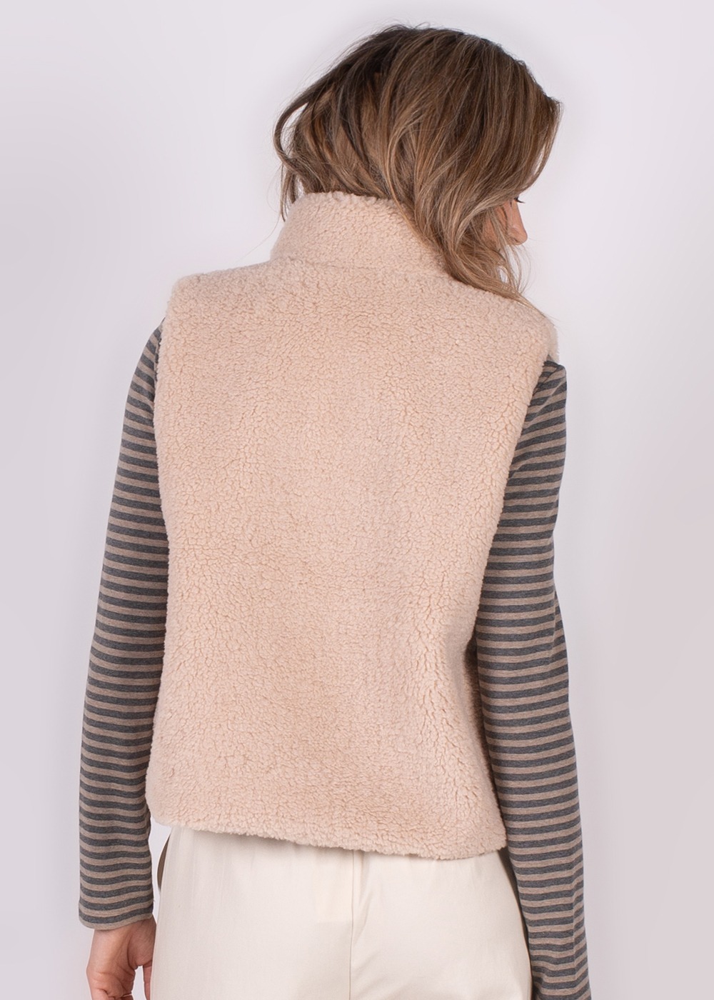 Achteraanzicht van een persoon in een zacht, lichtgekleurd teddy gilet, met daaronder een grijs gestreepte top en witte broek.