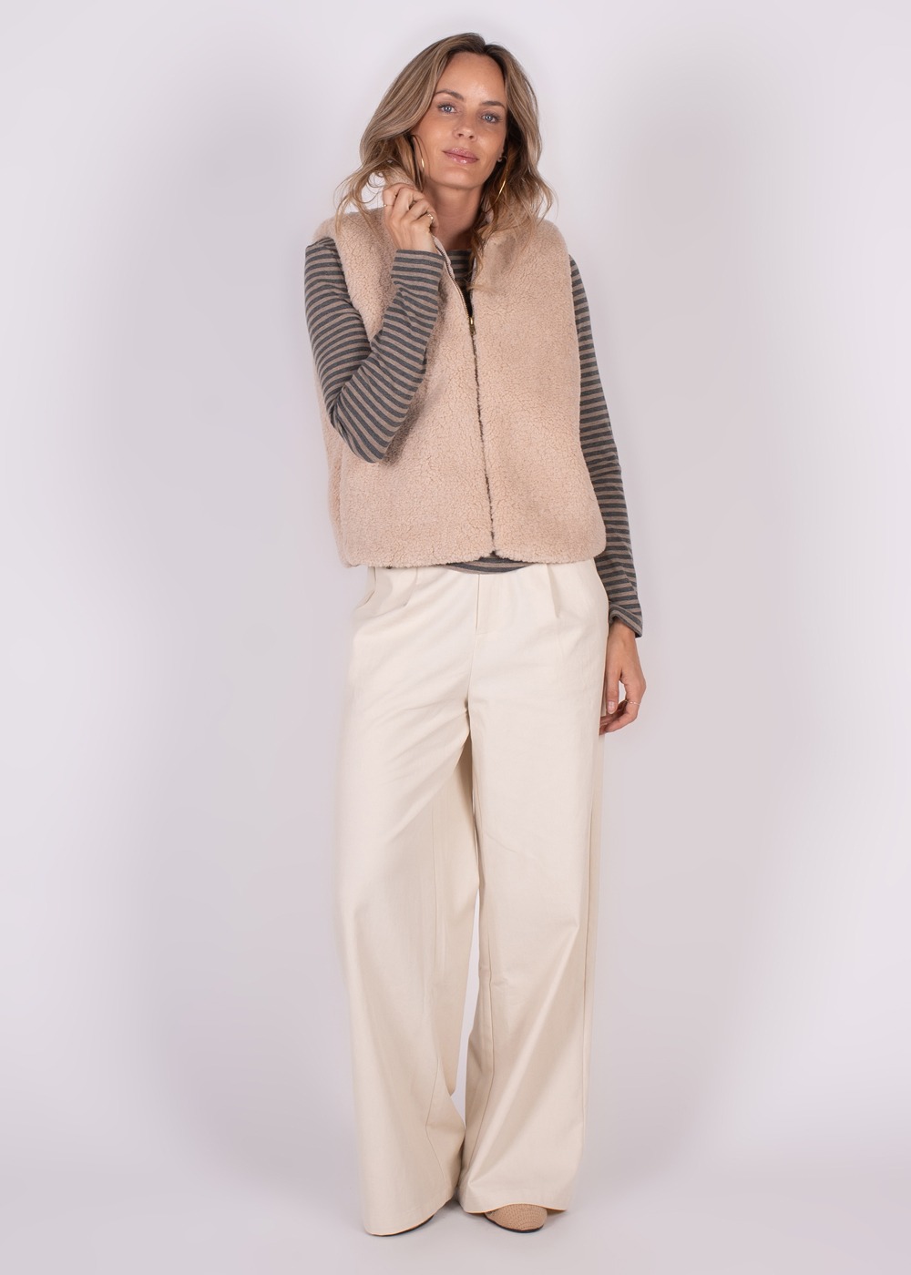 Een vrouw draagt een beige Regina gilet over een gestreept shirt met lange mouwen en wijde, crèmekleurige broek.
