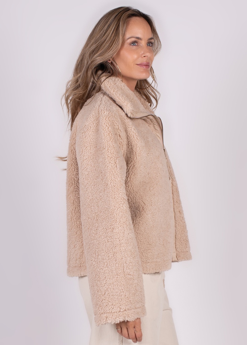 Een vrouw draagt een lichtbeige, pluche gilet met hoge kraag, gezien vanuit een zijaanzicht tegen een neutrale achtergrond.