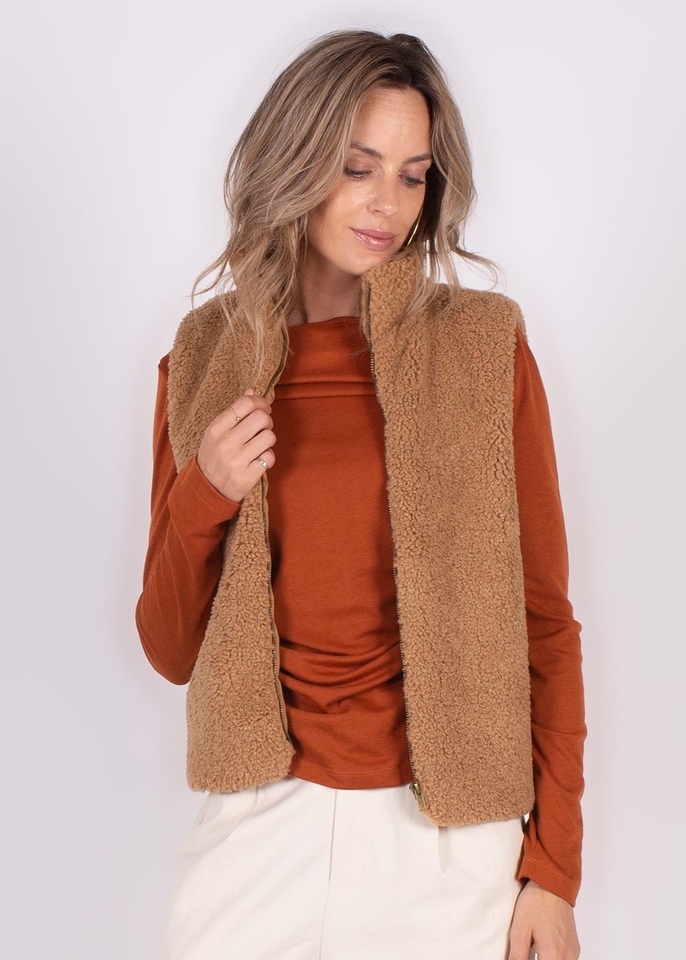 Model draagt een bruine teddy gilet over een roestkleurige trui met lichte broek tegen een neutrale achtergrond.