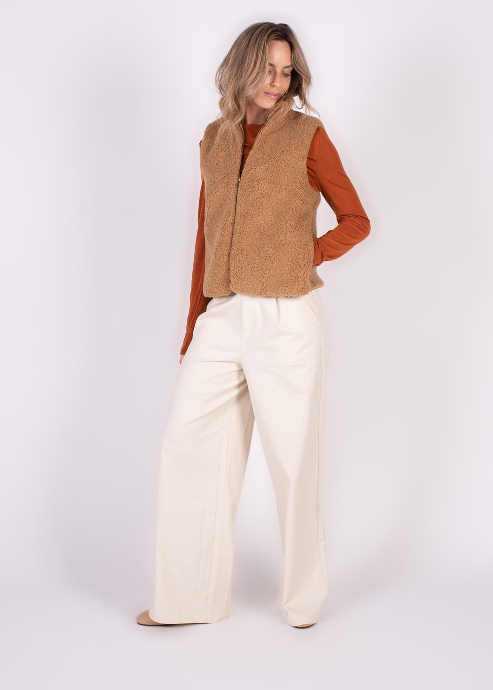 Model draagt een bruine teddy gilet over een oranje trui met wijde witte broek, tegen een neutrale achtergrond.