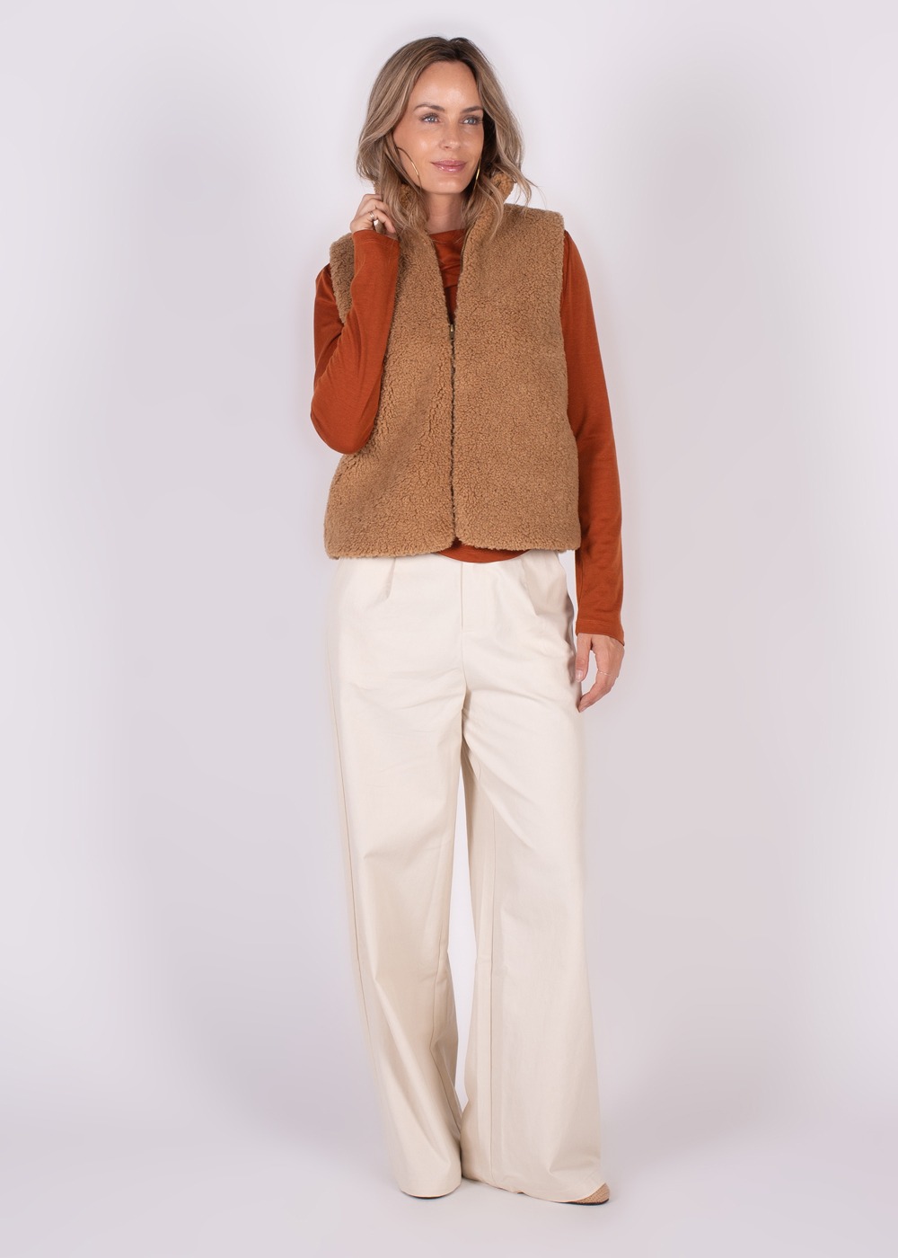 Model draagt een bruin teddy gilet en een roestkleurige trui met beige wijde broek, tegen een neutrale achtergrond.