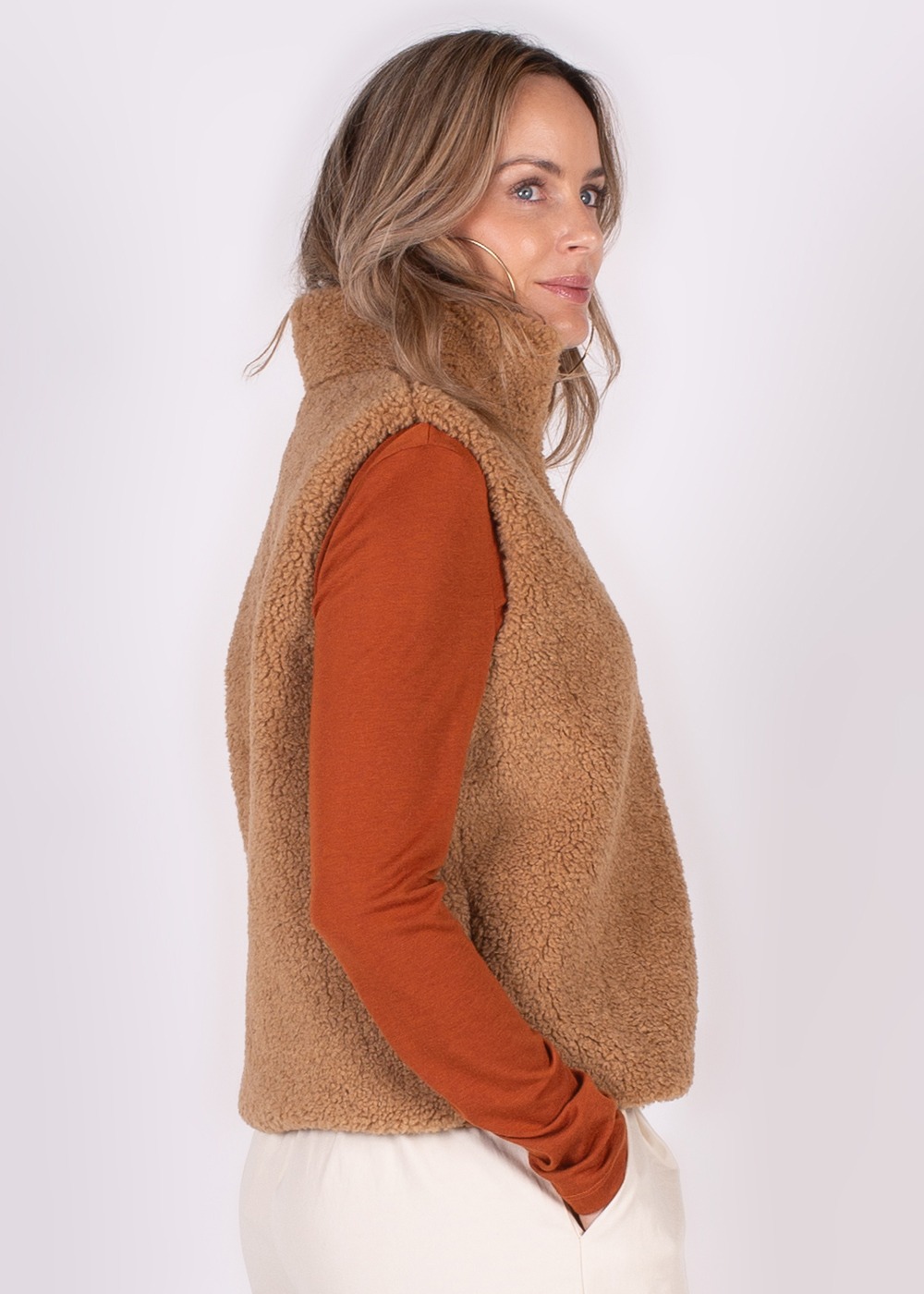 Model in zijaanzicht draagt een beige teddy gilet genaamd "Regina" over een roestkleurige trui.